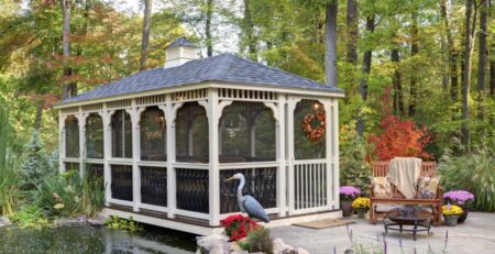 White vinyl rectangle gazebo with a cupola, next to a decorative pond with trees in background.
