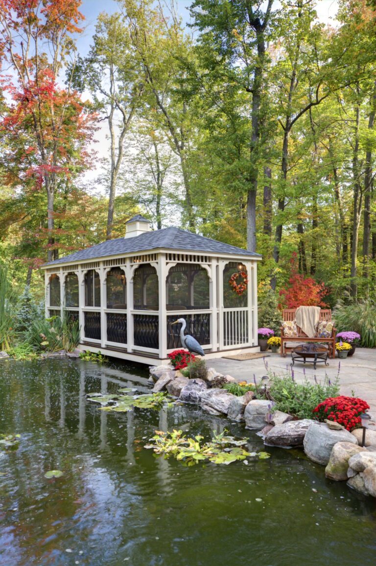 White vinyl rectangle gazebo with a cupola, next to a decorative pond with trees in background.