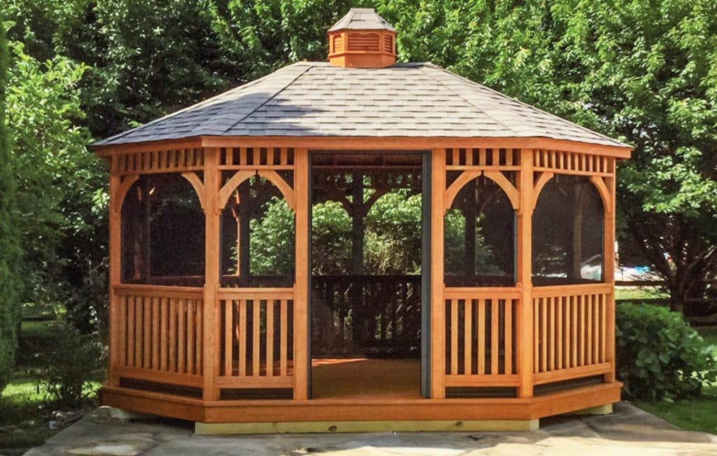 Charming wooden oval gazebo with a shingled roof, set in a lush garden.