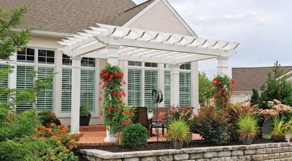 White vinyl pergola attached to a house with red flowering vines climbing the columns, surrounded by colorful landscaping including ornamental grasses, hydrangeas, and shrubs in a raised stone-walled garden bed