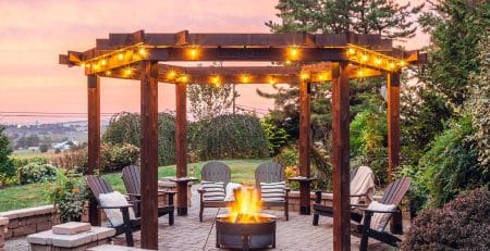 Country Lane Gazebos Ring of Fire cedar pergola with string lights, Adirondack chairs, and a fire pit on a paver patio at sunset