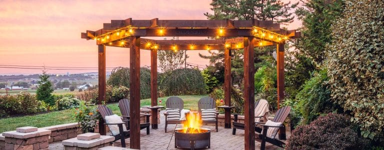 Country Lane Gazebos Ring of Fire cedar pergola with string lights, Adirondack chairs, and a fire pit on a paver patio at sunset