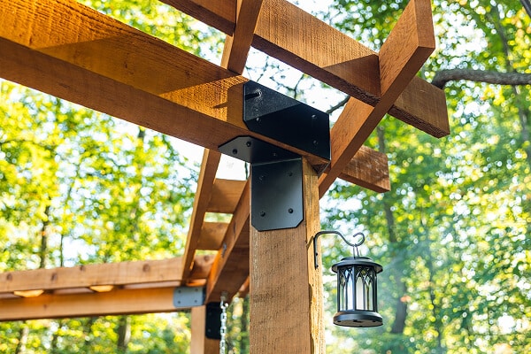 Close-up of a cedar pergola post with black steel bracket hardware, crossbeam joinery, and a hanging lantern surrounded by trees.

