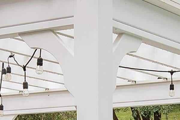 Close-up of a white vinyl pergola post with decorative braces, rafters, and string lights on a green lawn.

